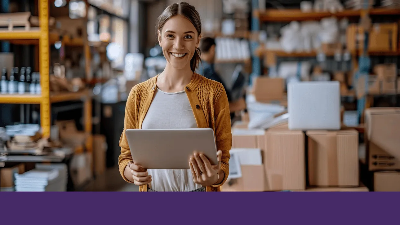 Smiling person in a warehouse setting, holding a tablet. Shelves with boxes and products are visible in the background